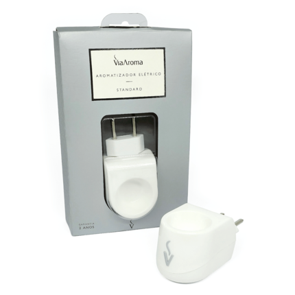 Aromatizador elétrico bivolt Via Aroma branco de tomada, feito de plástico, fora da embalagem, posicionado à direita da sua embalagem de papelão cinza, que possui uma janela transparente mostrando o encaixe do produto. Vendido pela loja Natureza Divina.