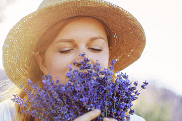 Mulher de chapéu de palha cheirando buquê de flores de lavanda com os olhos fechados.