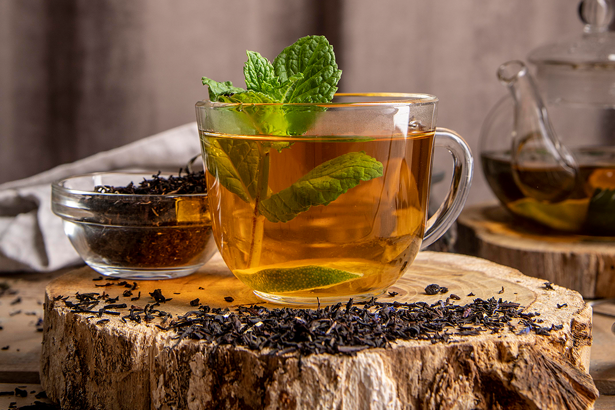 Freepik Xícara de vidro com chá-verde e folhas de hortelã, acompanhada de um recipiente com folhas secas e uma chaleira de vidro ao fundo.